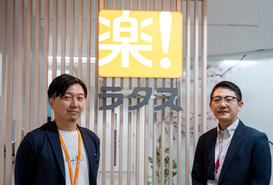 ラクスのロゴを背景に立っている2人の男性。